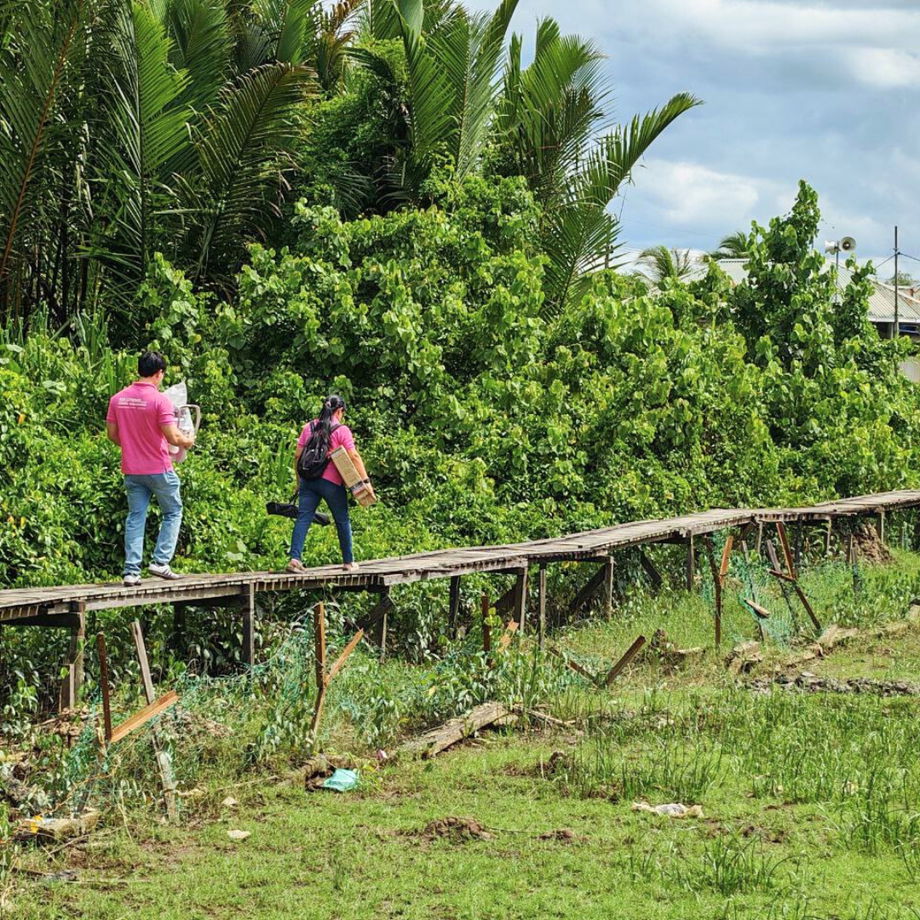 GHL VisionFirst team carrying optical equipment through muddy road and raised timber walkway at SK Muhibbah
